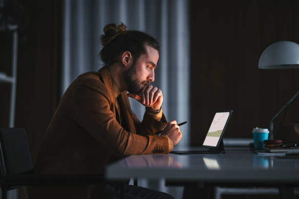 Pensive man working on laptop in office Side view of serious thoughtful adult bearded male in stylish wear sitting at table with modern laptop and analyzing business information while working in office in late evening worker working stock pictures, royalty-free photos & images