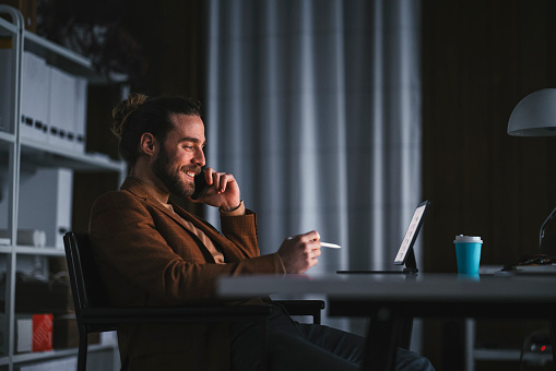 Side view of smiling adult bearded male in smart casual outfit talking on mobile phone while working with tablet and stylus pen in modern workplace
