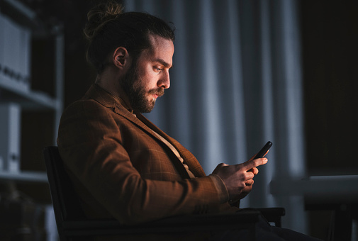 Side view of focused adult bearded male in stylish outfit checking messages on mobile phone while working in dark office at night