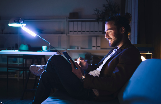 Side view smiling young male in smart casual wear using tablet and stylus while sitting with crossed legs on cozy couch in dark room