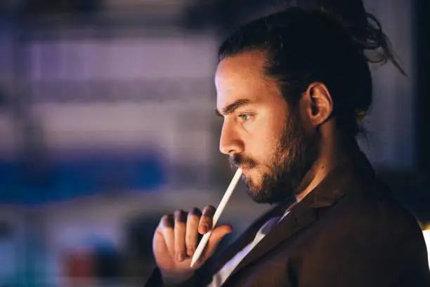 Side view thoughtful bearded male in black jacket touching lips with stylus while working in dark room