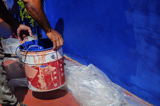 Close-up shot of an unrecognizable Middle Eastern Man lifting the paint can as he repainting the exterior wall of his new house with blue color. The floor is covered with plastic to protect it from the paint drop.