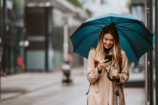bella ragazza con un ombrello in piedi sotto la pioggia che digita un messaggio. - ombrello foto e immagini stock