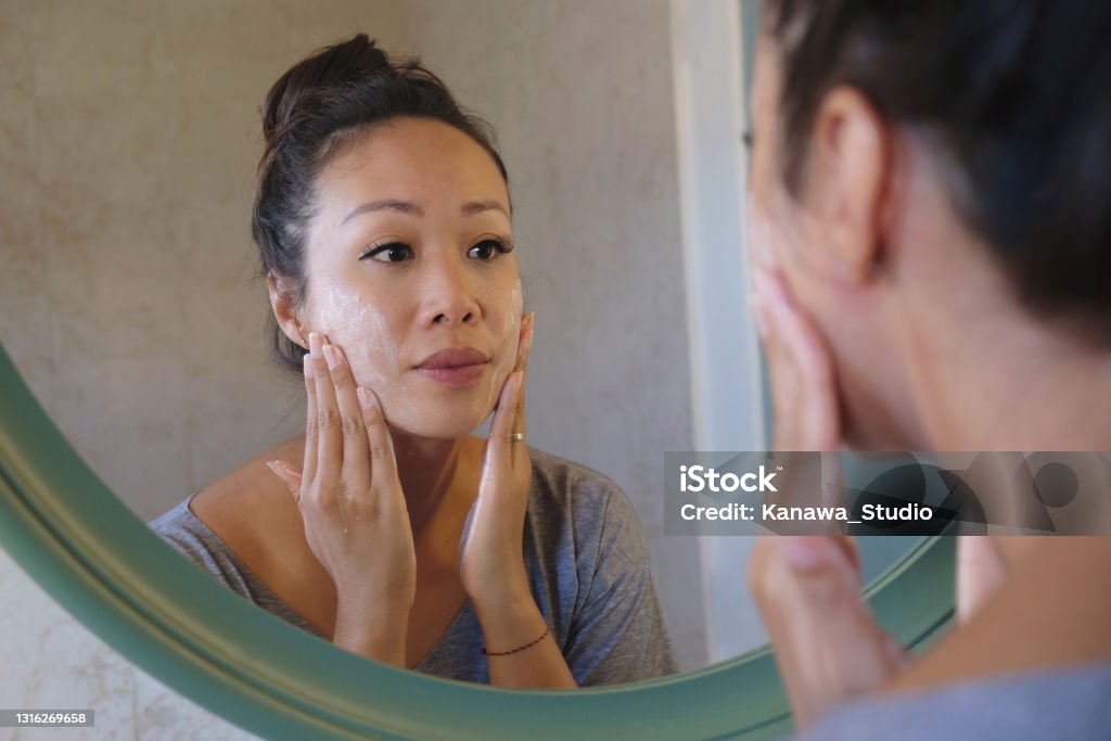 Indonesian Woman Washing Her Face Using Beauty Cleanser Soap Rear-view shot of a beautiful Indonesian woman washing her face using beauty cleanser soap. She's looking at her reflection in the mirror while thoroughly rubbing her face. Exfoliation Stock Photo
