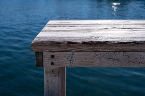 Old empty grey wooden bench, pier over blur blue sea background. Waterfront fish tavern table, wood planks, platform for relaxation, sunny summer day. Vacation, travel, tourism at nature.