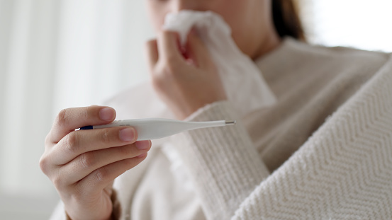 Sick young asian woman sit under blanket on sofa and sneeze with tissue paper at home. Female blow nose, coughing, sneezing, check temp in tissue at home, suffering from flu. Cold and fever concept