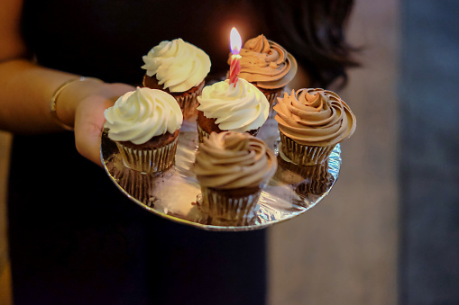 Close-up shot of an unrecognizable Asian woman carrying a tray of homemade cupcakes with a lighted candle on one of them to surprise her friends birthday.