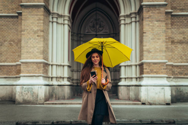 istock Young beautiful woman using a smartphone and holding a yellow umbrella 1316280765