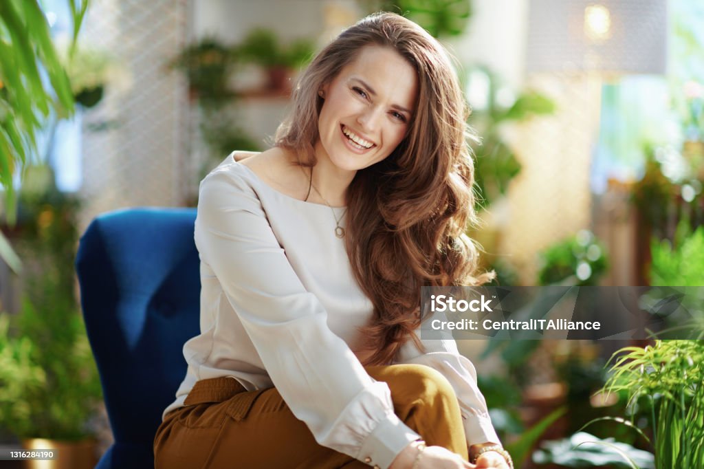 smiling trendy woman with long wavy hair in house in sunny day Green Home. Portrait of smiling trendy middle aged woman with long wavy hair in the modern house in sunny day in green pants and grey blouse sitting in a blue armchair. One Woman Only Stock Photo