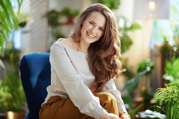 smiling trendy woman with long wavy hair in house in sunny day Green Home. Portrait of smiling trendy middle aged woman with long wavy hair in the modern house in sunny day in green pants and grey blouse sitting in a blue armchair. 40 44 years stock pictures, royalty-free photos & images