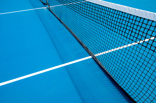 A tennis court net casts a shadow on the blue surface of the court.