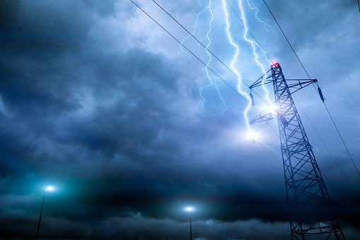 Thunderstorm lightning hitting powerline tower with flashes and plasma balls forming along electricity cables. Possible damage or blackout. Cloudy sky with stormy weather at dusk. Digital composite.
