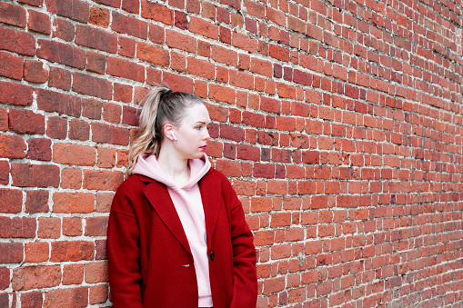 Young woman wearing earbuds, woolen coat and hoodie is standing by the brick wall and is looking away. Copy space