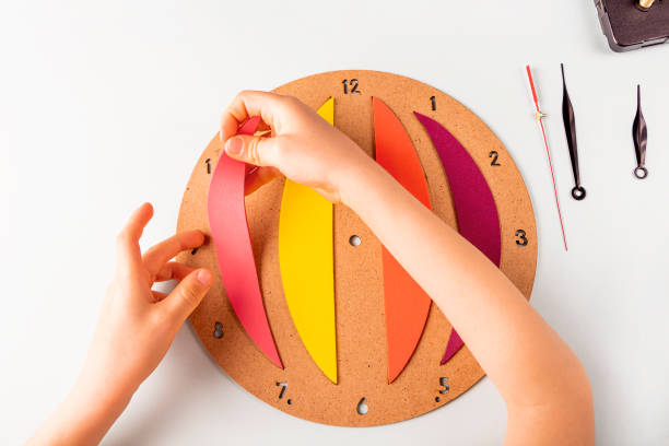 istock DIY and kid's creativity. Step by step instruction: how to wall clock. Step3 childs hands glue colored parts to wood pannel. Children Craft workshop. 1316306689