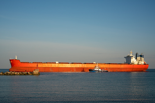 Large ocean-going cargo ship in orange. The dry cargo ship enters the port using a tug