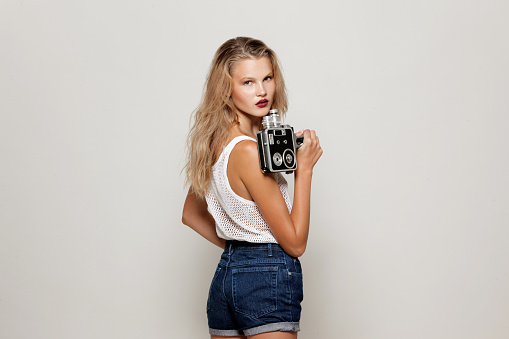 Portrait of girl posing in white fashion clothes in studio, holding a retro camera in hand, looking away.