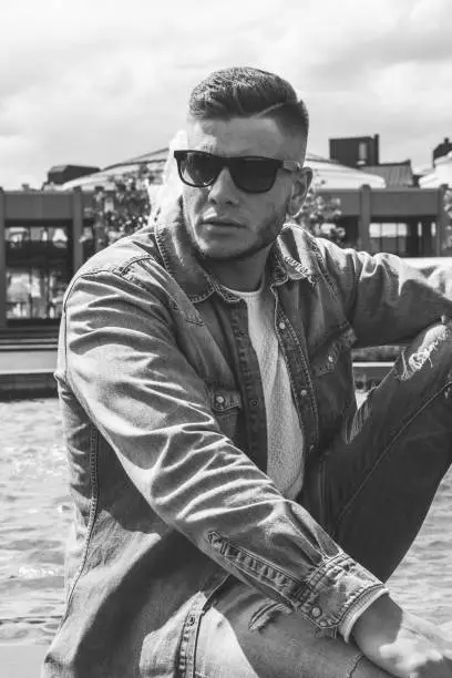 Trendy handsome young man wearing sunglasses seated alongside a canal looking thoughtfully off to the side in a close up greyscale portrait