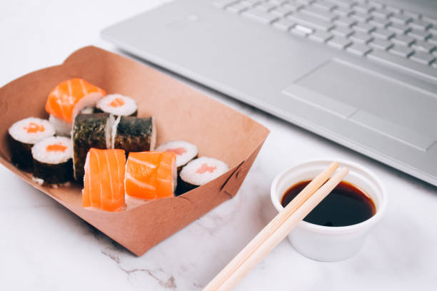 istock Different sushi rolls with salmon in delivery paper box on white marble table background with laptop - futomaki, uramaki and hosomaki. Order food online and home delivery food concept 1316322998