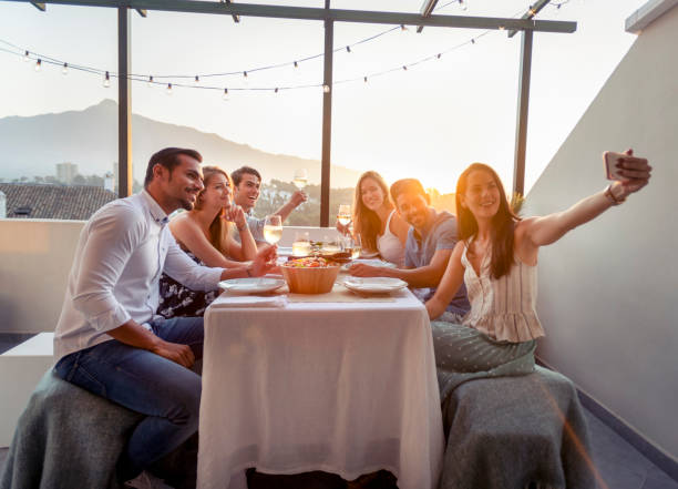 Group of friends taking a selfie or a video call at a dinner party with a mobile phone Group of friends taking a selfie or a video call at a dinner party with a mobile phone. Time is sunset and they are drinking wine sitting at the dinner table. There is healthy food on the table. There is a nice view from the patio. Everybody is happy and smiling. exclusive dinner stock pictures, royalty-free photos & images
