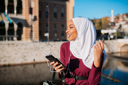 Happy young woman of middle eastern ethnicity enjoying the sunny day outdoors and smiling, using a smart phone to listen to music