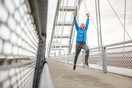 Ecstatic senior male athlete running on a bridge and celebrating while jumping in the air with hands up.