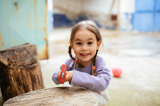 Little girl aged 4 hammering a nail to the wooden cabin wall. The smiling girl is practicing the ability to correctly hammer a nail.