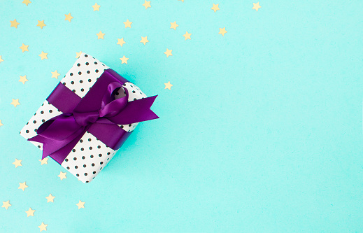 A gift box wrapped in polka dot paper and a purple ribbon, with gold stars on a pale turquoise background.  \nShot from above. \nBirthday/ Celebration/ Shopping background.