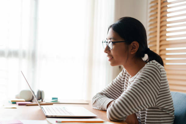 istock Side view. Happy asian girl wear glasses looking screen computer while sitting learning online on laptop at home.  Concept of online learning at home 1316365588