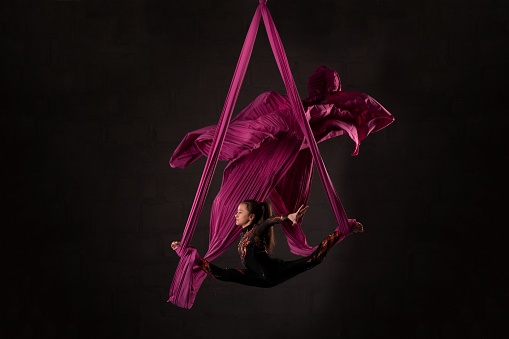 Full body side view of flexible young female aerial dancer doing splits on hanging ribbons against black background