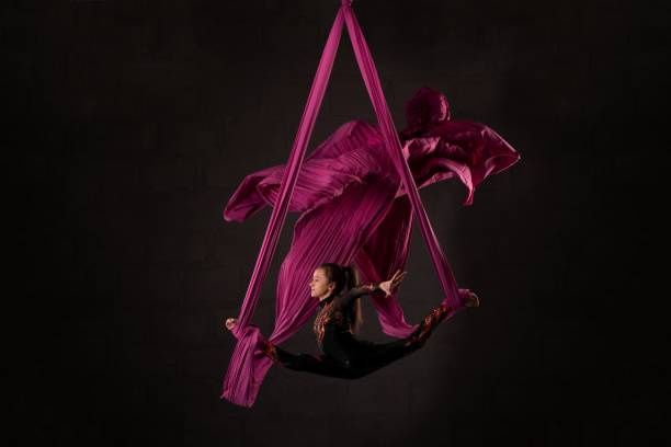 Woman performing acrobatic dance on aerial silks Full body side view of flexible young female aerial dancer doing splits on hanging ribbons against black background acrobatic activity stock pictures, royalty-free photos & images