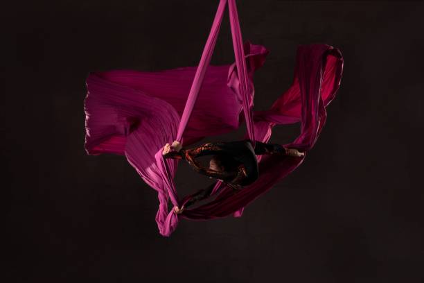 Teen girl performing acrobatic split over head on aerial silks Full body side view of flexible teen girl doing splits over head on hanging ribbons aerial silk against black background acrobatic activity stock pictures, royalty-free photos & images