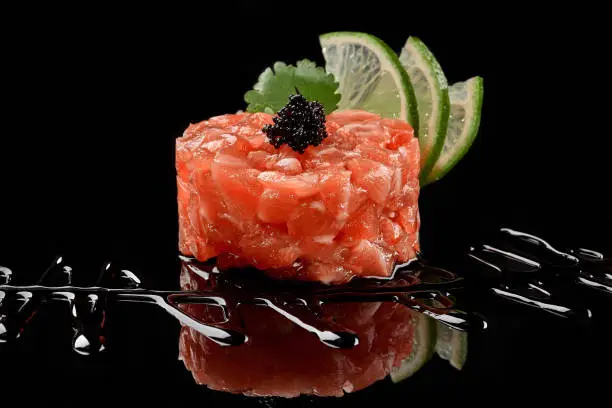 Closeup of raw salmon tartare garnished with caviar, lime slices, fresh parsley leaf and soy sauce on black background with reflection