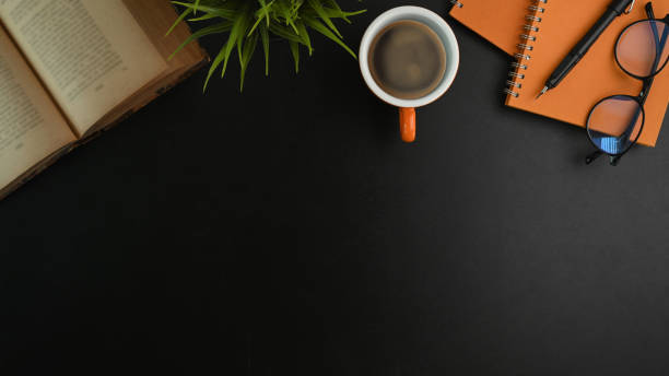 Top view office workspace with book, coffee cup, notebook and plant on black table.