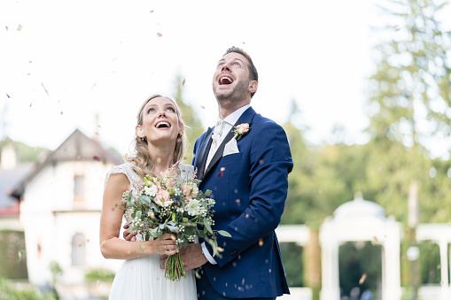 beautiful newly married wedding couple together outdoors on bright sunny summer day with flower pedals falling over them, shallow focus, background blurred