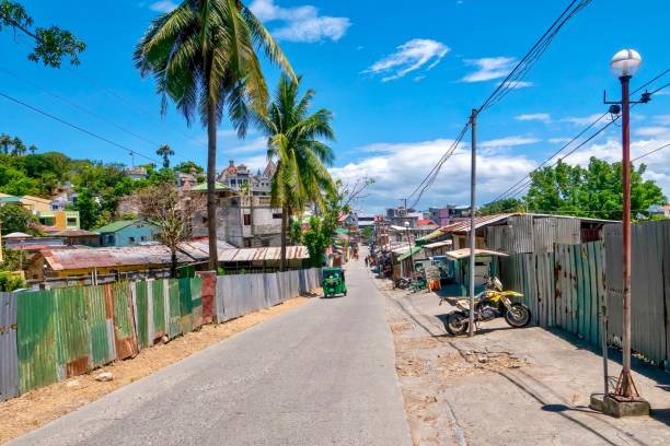 A road leading to the popular resort town of Sabang Beach, Puerto Galera, Philippines. Sabang Village, Puerto Galera, Philippines - May 4, 2021: A narrow road lined with corrugated metal sheeting, palm trees, and simple buildings on the outskirts of a popular dive resort town on Mindoro Island. philippines tricycle stock pictures, royalty-free photos & images