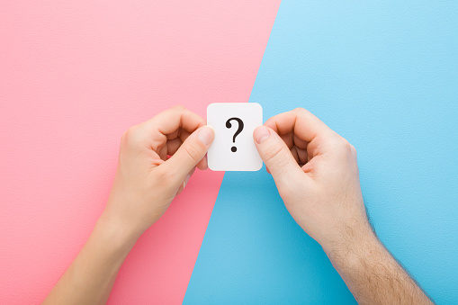 Young adult man and woman hands together holding white card of question mark on light blue pink table background. Pastel colors. Concept of couple relationship issues. Closeup. Top down view.