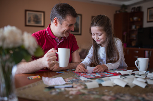 Man smiling and having a cup of coffee when his daughter fills in UEFA sticker album