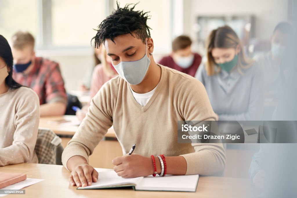 Black college student with face mask taking notes during lecture in the classroom. African American student writing in notebook while wearing face mask and attending lecture at the university during coronavirus pandemic. Protective Face Mask Stock Photo