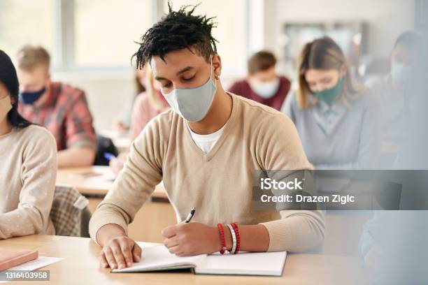 Black College Student With Face Mask Taking Notes During Lecture In The Classroom Stock Photo - Download Image Now