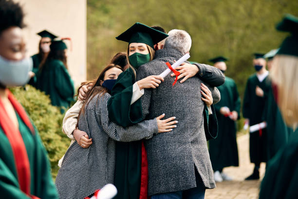 Happy student embracing her family on her graduation day during COVID-19 pandemic. Happy female graduate embracing with her family after receiving a diploma during coronavirus pandemic. ritual mask stock pictures, royalty-free photos & images