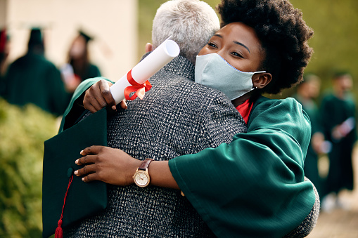 Happy African American student with face mask embracing her father on her graduation day.