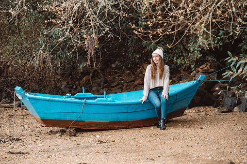 Female in hat and sweater sitting on old wooden boat moored on sandy shore in autumn and looking away