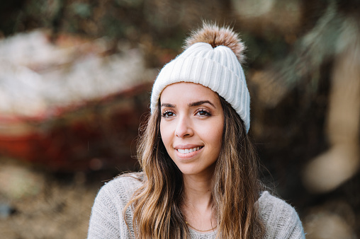 Cheerful young female traveler in warm knitted hat and sweater smiling and looking away while resting on blurred coast with old boat