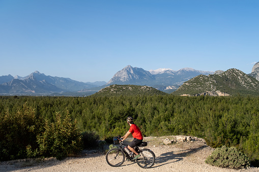 Cyclist in red shirt with a helmet, 25-30 years old. .He's riding a bicycle. At the same time, he is looking at the landscape with his head turned upside down. A dense forest is behind him, the mountains. An exquisite nature view is open air. Early morning hours