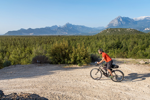 Rider, 25-30 years old, in red shirt and helmet. .He's riding a bicycle. . there is a dense forest and mountains. An exquisite nature view is open air. Early morning hours.Applying force to pedal.In motion