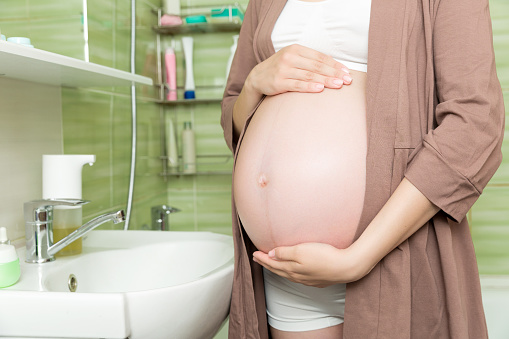 Young pregnant woman dressed in a robe in the toilet at home. Cute adult girl in the third trimester of pregnancy in a home bathroom. Pregnancy, maternity and bodycare concept