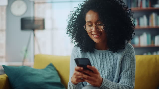 Portrait of a Beautiful Authentic Latina Female in a Stylish Cozy Living Room Using Smartphone at Home. She's Browsing the Internet and Checking Videos on Social Networks and Having Fun.