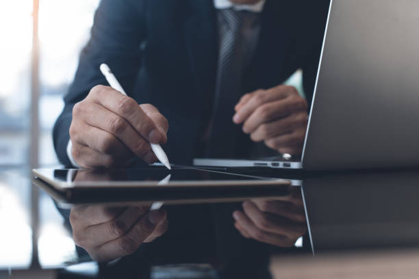 Businessman working in modern office Businessman in black suit working in modern office. Business man with stylus pen signing on digital tablet screen, reviewing business report with laptop computer on glass table, close up contracting stock pictures, royalty-free photos & images