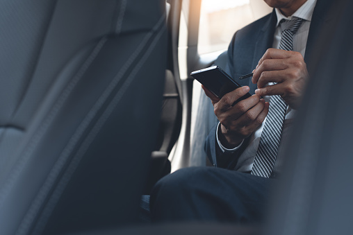 Asian businessman in black suit, executive manager using mobile smart phone taking note via application notepad inside a car on backseat. Business man going to workplace by taxi, portable office, business concept