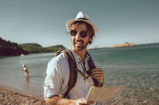 Smiling tourist man standing with map and backpack near the sea.